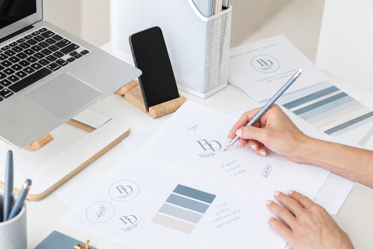 A close-up of a person's hands writing on branded design concept sheets with blue-gray and beige color swatches, set on a white desk alongside a laptop, smartphone, and office accessories.