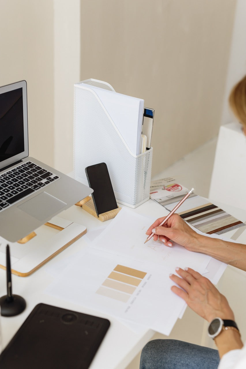 A person at a desk writing on a paper with color swatches, surrounded by a laptop, phone, and white organizer in a bright indoor workspace.